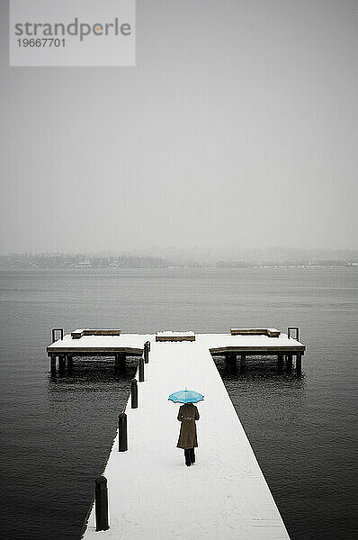 Eine junge Frau geht auf einem schneebedeckten Steg mit Blick auf einen See und hält einen leuchtend blauen Regenschirm in der Hand.