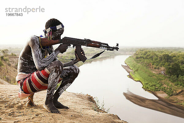 Ein junger Mann hält sein Kalaschnikow-Gewehr in der Hand  während er den Omo-Fluss im abgelegenen Omo-Tal in Äthiopien überblickt