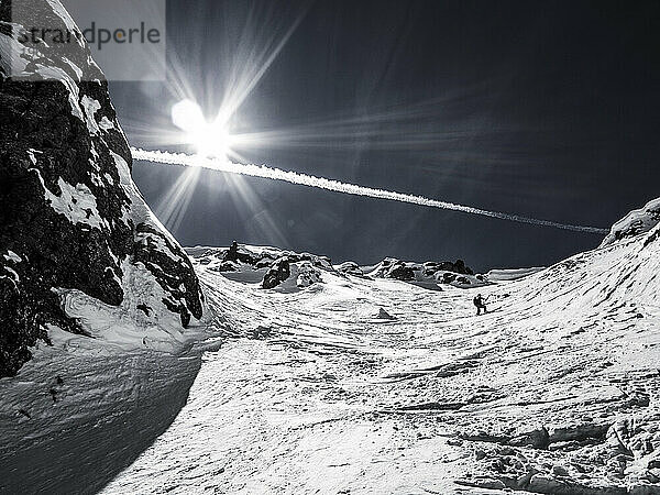 Ein Mann  der im Hinterland Ski fährt  mit Sonneneruption und Jet-Kondensstreifen am Himmel.