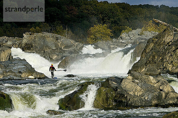 Ein Mann steht beim Stand-Up-Paddleboarden am Fuße einer großen Wasserfallgruppe.