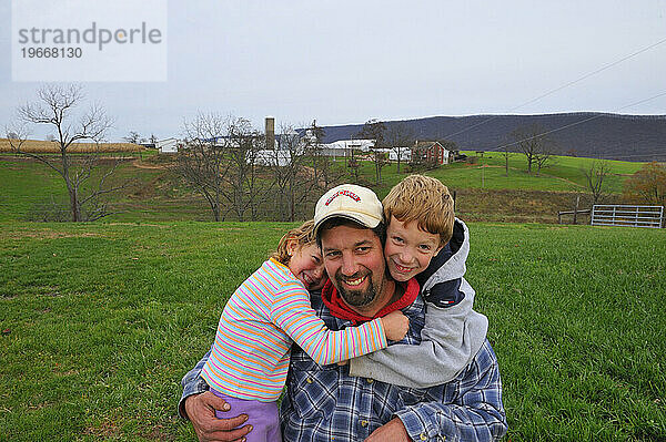 Ein Maisbauer posiert mit seinen beiden lächelnden Kindern für ein Porträt auf einer riesigen grünen Wiese auf seiner Farm in Bloomsburg  PA.