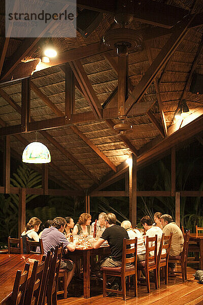 Eine Gruppe Urlauber isst an einem Tisch unter einer Holzkonstruktion in einer Öko-Lodge in einem fremden Land.