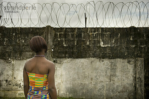 Eine junge liberianische Frau in einem farbenfrohen afrikanischen Kleid steht vor einer großen Mauer und starrt auf den Stacheldraht oben.