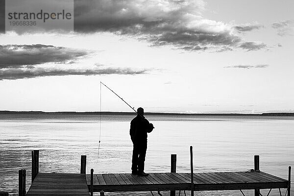 Ein Schwarz-Weiß-Bild eines Mannes  der sich als Silhouette vor einem dramatischen Himmel abzeichnet und eine Angelrute in der Hand hält.