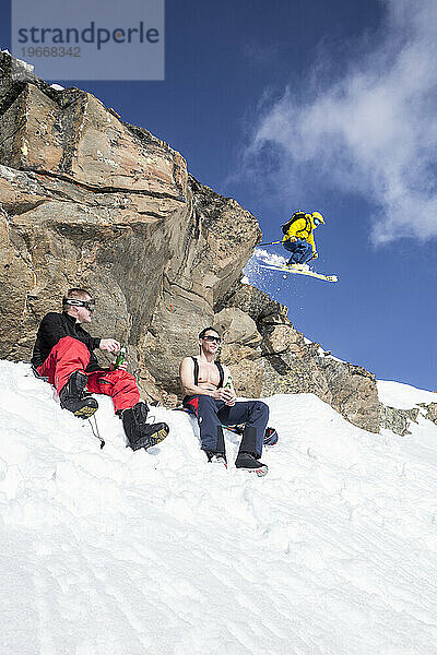 Skifahrer springt über zwei Personen  die unter einer Klippe etwas trinken