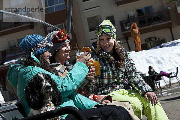 Drei Freunde (ein Mann und zwei Frauen) hängen mit Bier und einem Hund vor einem Skigebiet herum.