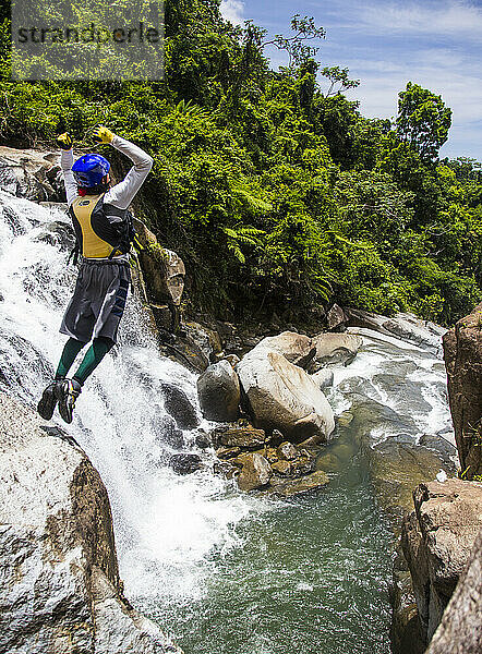 Ein Mann macht einen Backflip von einem Felsvorsprung in der Nähe eines Wasserfalls an einem Fluss in Puerto Rico.