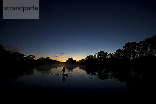 Silhouette eines Mannes beim Stand-Up-Paddleboarden im Sonnenaufgangslicht mit Bäumen  die sich im Wasser spiegeln.