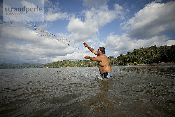 Ein großer Mann steht hüfthoch im Wasser und wirft unter blauem Himmel ein Netz in das ruhige Wasser.