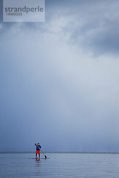 Ein Mann beim Stand-Up-Paddleboarden auf einem Bergsee im Winter  während ein Sturm aufzieht.