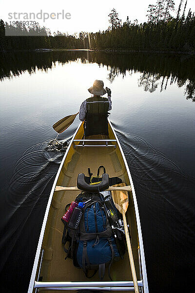 Eine Frau paddelt mit ihrem Kanu  während die Sonne über den Bäumen am Horizont der Boundary Waters in Minnesota aufgeht.