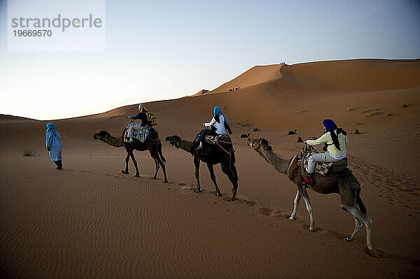 Wüstenkamelführer nehmen Touristen bei Sonnenaufgang mit auf einen Ausritt über die Sanddünen.