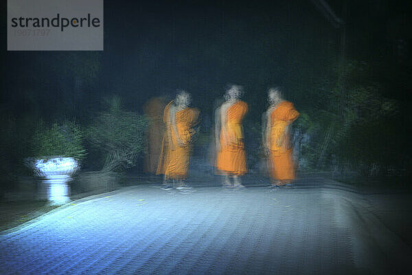 Drei buddhistische Mönche stehen nachts in einem Innenhof  der nur von einer Straßenlaterne beleuchtet wird. Chiang Rai  Thailand. (Bewegungsunschärfe)