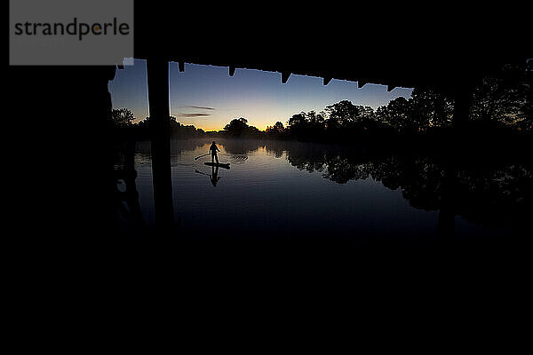 Silhouette eines Mannes beim Stand-Up-Paddleboarden im Sonnenaufgangslicht mit Bäumen  die sich im Wasser spiegeln.