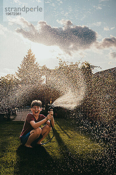 Fröhlicher Junge  der an einem Sommertag Wasser aus einem Schlauch in einem Garten versprüht.