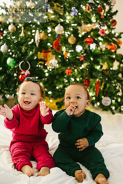 Baby twin siblings together laughing in front of Christmas tree