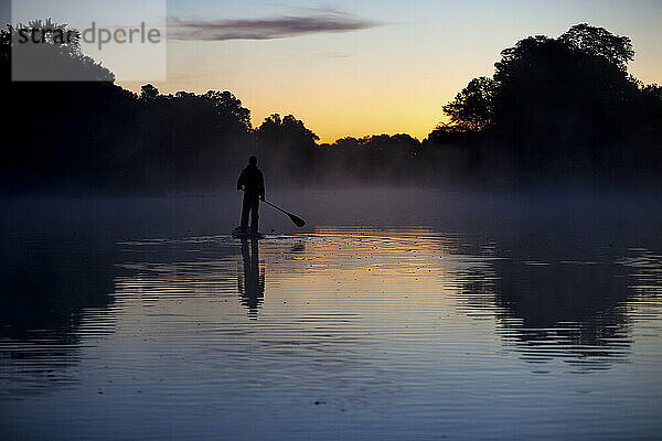 Silhouette eines Mannes beim Stand-Up-Paddleboarden im Sonnenaufgangslicht mit Bäumen  die sich im Wasser spiegeln.