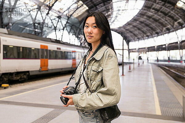 Schönes Mädchen hält eine Kamera in einem Bahnhof und blickt in die Kamera.