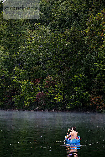 Ein Mann schwimmt an einem ruhigen  regnerischen Tag in einem Kajak auf einem See mit grünem  üppigem Hintergrund.