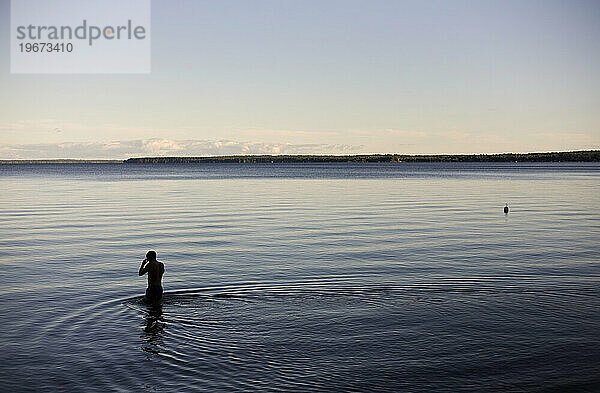 Ein Mann  dessen Silhouette sich vor dem ruhigen Wasser abzeichnet  watet in der Abenddämmerung in dunkles  blaues Wasser.