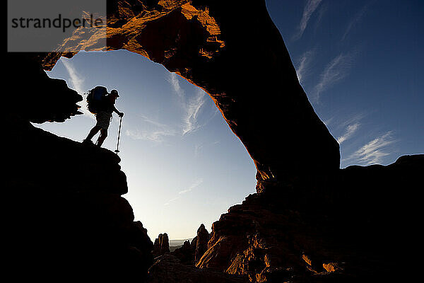 Silhouette eines Wanderers  der unter einem natürlichen Felsbogen im Arches-Nationalpark in Utah steht.