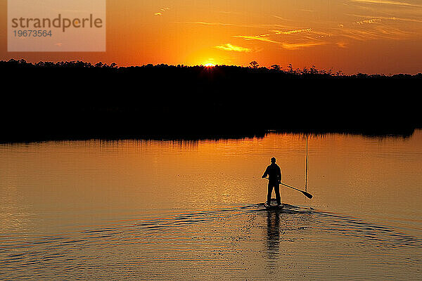 Silhouette eines Mannes beim Stand-up-Paddeln im Abendlicht mit Bäumen  die sich im Wasser spiegeln.