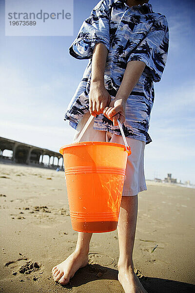 Little boy holds an orange sandbucket at the beach.