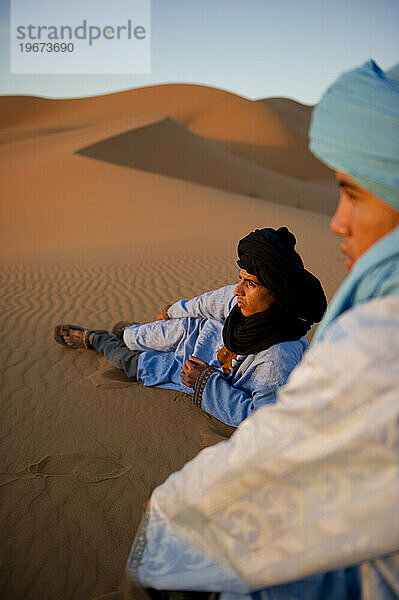 Two boys in traditional North African dress sit on the top of a sand dune at sunrise.