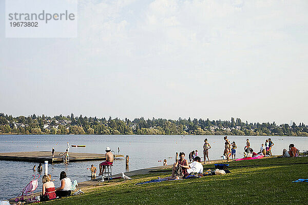Menschen sitzen in der Sonne am Ufer eines Sees  während sich im Hintergrund Menschen sonnen und schwimmen.