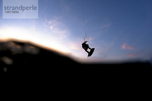 Zweistufige Ansicht eines Mannes beim Kitesurfen und Fliegen in der Luft gegen Wasser und einem Sonnenuntergang.