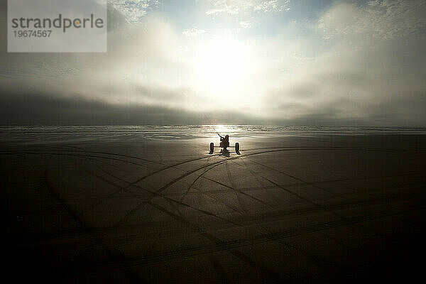 Silhouette eines männlichen Drachenbuggys  der in bedrohlichen Wolken und ungewöhnlichem Licht mit dunklen Spuren im Sand den Strand hinunterrollt.