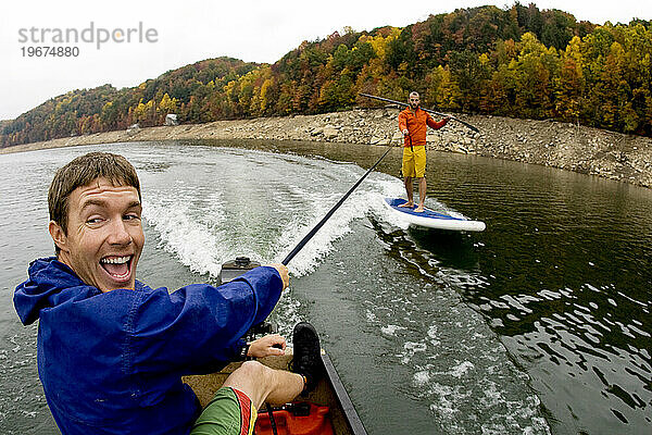 Zwei Männer surfen hinter einem kleinen Boot auf einem Stand-Up-Paddleboard.