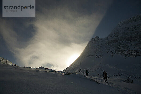 Silhouette zweier Backcountry-Skifahrer  die bei Sonnenaufgang einen Hang in den Bergen erklimmen.