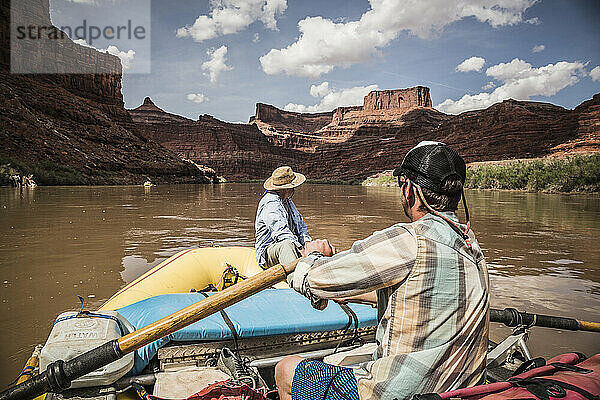 Ein Mann rudert mit einem Floß einen Fluss hinunter in einer Wüstenlandschaft.