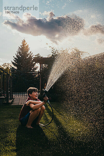 Fröhlicher Junge  der an einem Sommertag Wasser aus einem Schlauch in einem Garten versprüht.