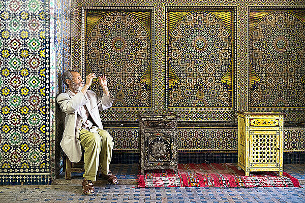 Ein vornehm aussehender Mann reinigt seine Brille  während er vor einer spektakulär gemusterten Mosaikwand sitzt