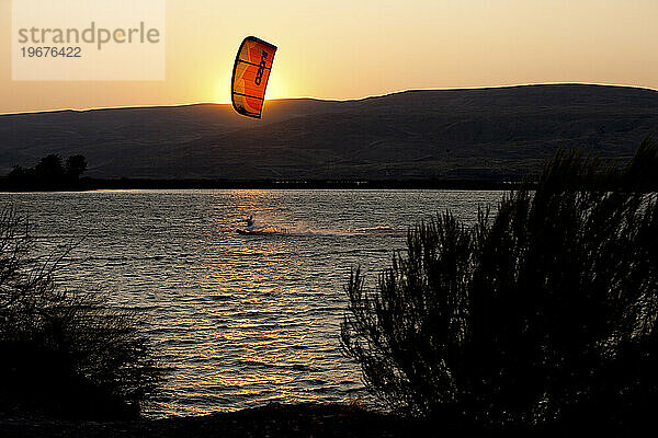 Die Silhouette eines männlichen Kiteboarders mit Sonnenuntergang  Wüstenbergen und Gestrüpp.