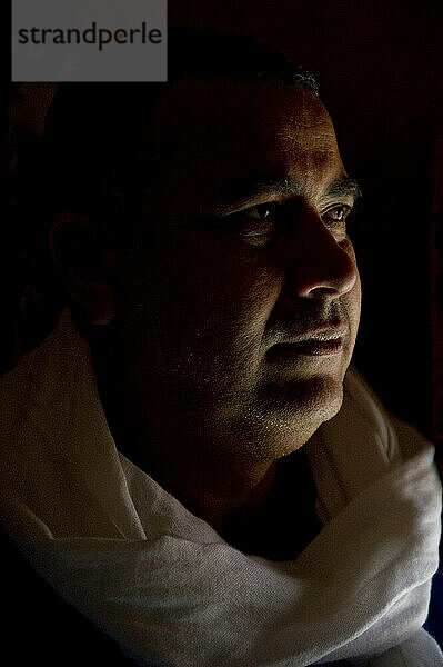 A Moroccan man stands in a dark room near a small window.