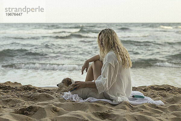 Glückliche junge Frau mit lockigem Haar hält Hund  während sie am Strand sitzt