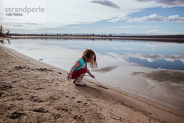 Junges Mädchen spielt mit Stock am Rande eines ruhigen Sees