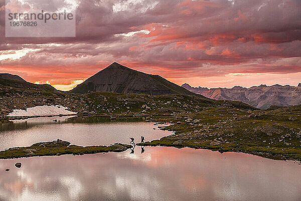 Zwei Menschen wandern bei Sonnenuntergang am Rande eines Sees in den Bergen entlang.