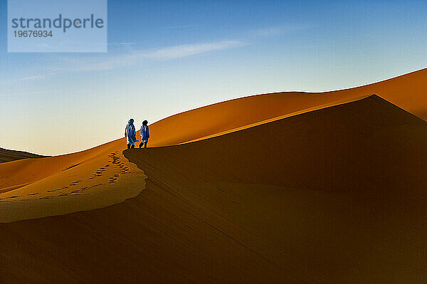 Zwei Jungen in traditioneller nordafrikanischer Kleidung stehen bei Sonnenaufgang auf der Spitze einer Sanddüne.