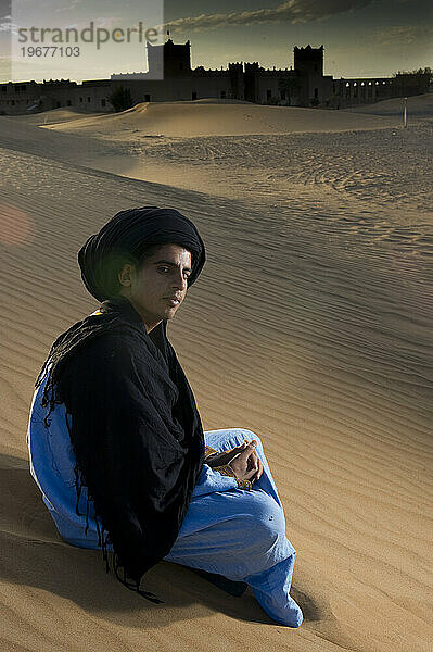 Ein Wüstenführer in traditioneller Kleidung sitzt auf einer Düne in der Nähe einer alten Wüstenoase.