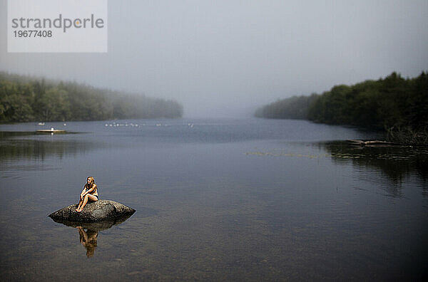 Ein junges Mädchen sitzt an einem nebligen Tag auf einem Felsen in einem kleinen Teich.
