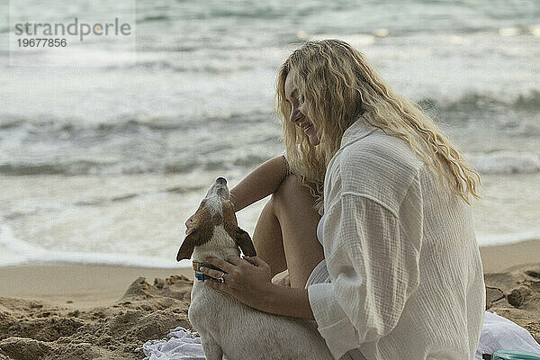 Glückliche junge Frau mit lockigem Haar hält Hund  während sie am Strand sitzt