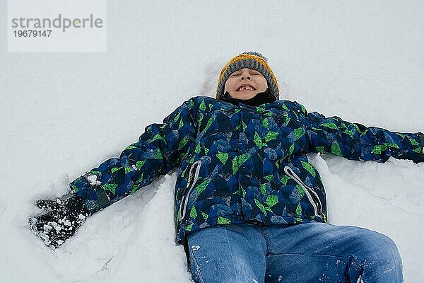 Fröhlicher Junge liegt im Schnee. Aktive Freizeit im Freien für Kinder in der Natur im verschneiten Wintertag
