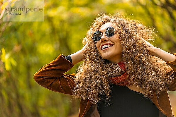 Glückliches Modell mit lockigem Haar und Sonnenbrille posiert und lächelt in einem Park im Herbst