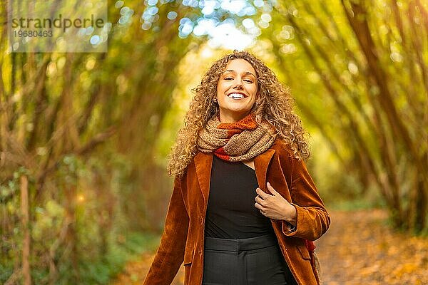Glückliche Frau in lässiger warmer Kleidung  die durch einen Park mit gefallenen Blättern im Herbst spaziert