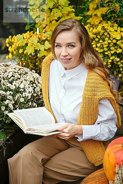 Draufsicht auf eine Frau mit einem Buch  die auf einer Bank zwischen Herbstblumen sitzt
