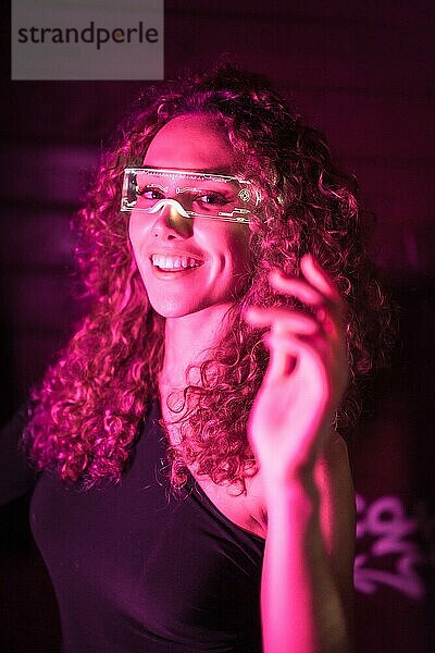 Vertikale Nahaufnahme einer glücklichen Frau  die eine intelligente Brille trägt und in die Kamera lächelt  mit rosa Neonlicht
